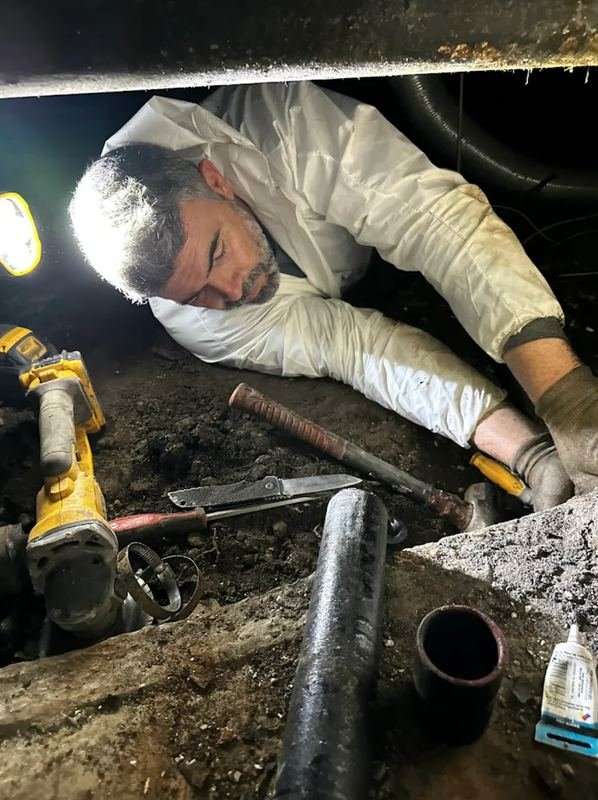 Plumber lying in a crawlspace cutting out old cast iron drain pipe